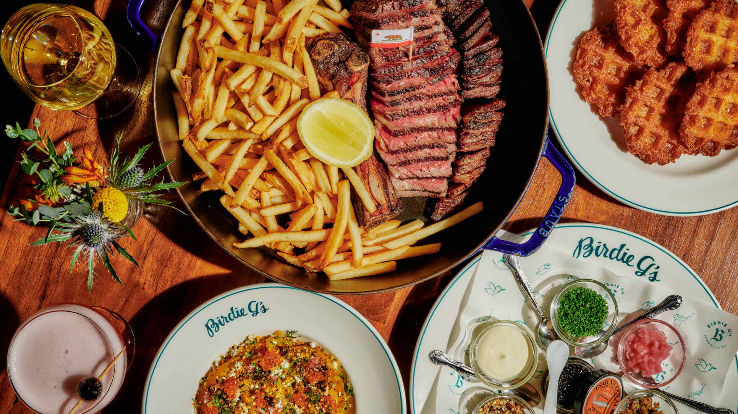 a variety of Birdie G's staple dishes on a wooden table, featuring our beet salad, prime steak frites, potato waffles with accoutrements and a refreshing cocktail